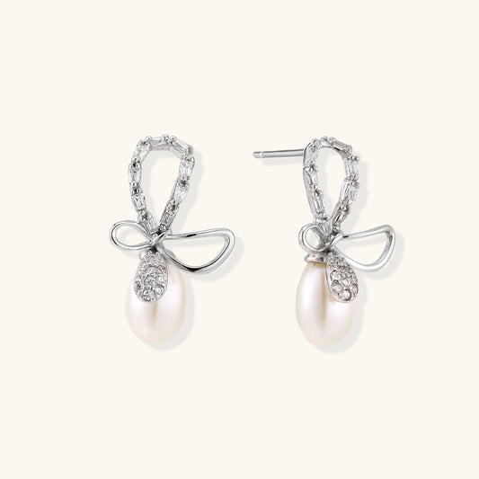 Silver bow-shaped stud earrings with baguette crystals and dangling white freshwater pearls on cream background